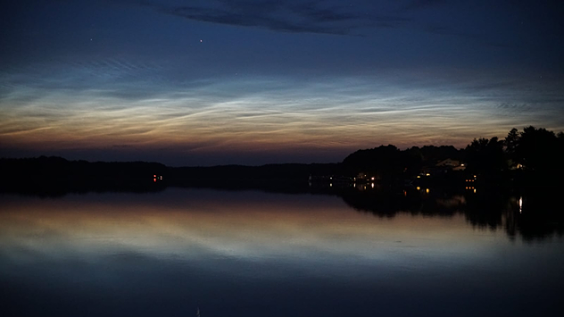 Nubes nocturnas luminosas sobre un lago al atardecer. El cielo brilla con tonos azules y naranjas, y el agua refleja la escena.