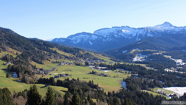 Sonnige Landschaft im Bregenzerwald mit grünen Wiesen, verstreuten Dörfern und schneebedeckten Alpen im Hintergrund.