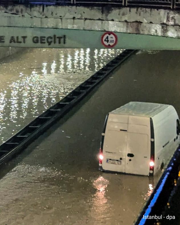Una furgoneta blanca está aparcada en una calle inundada, debajo de un puente en Estambul. El agua llega hasta las ruedas y refleja las luces del vehículo.