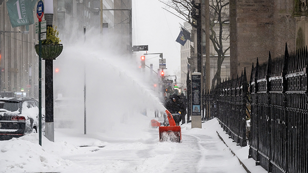 A person clears a snow-covered sidewalk in a city center with a snow blower during heavy snowfall.