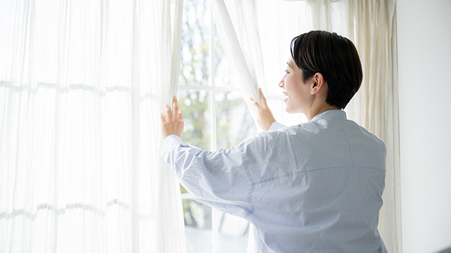 Frau öffnet Vorhänge und Fenster, um den Raum zu lüften. Helles Sonnenlicht scheint ins Zimmer. Symbolbild für Lüften bei Pollenallergie.