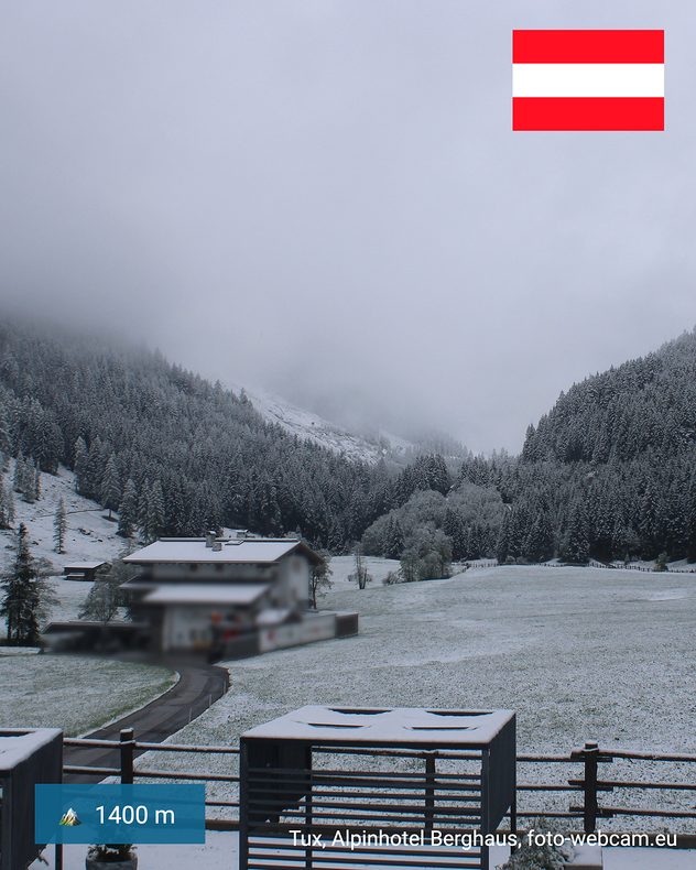 Webcambeelden uit Tux in Tirol, op 1400 meter hoogte. Besneeuwde weiden, huizen en dichte bergbossen liggen onder diepe wolken.