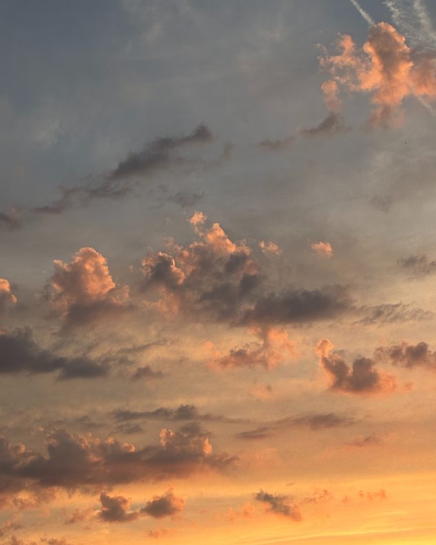 Scattered cumulus clouds glowing orange and peach at sunset beneath a hazy gray sky, with warm golden light intensifying toward the horizon.