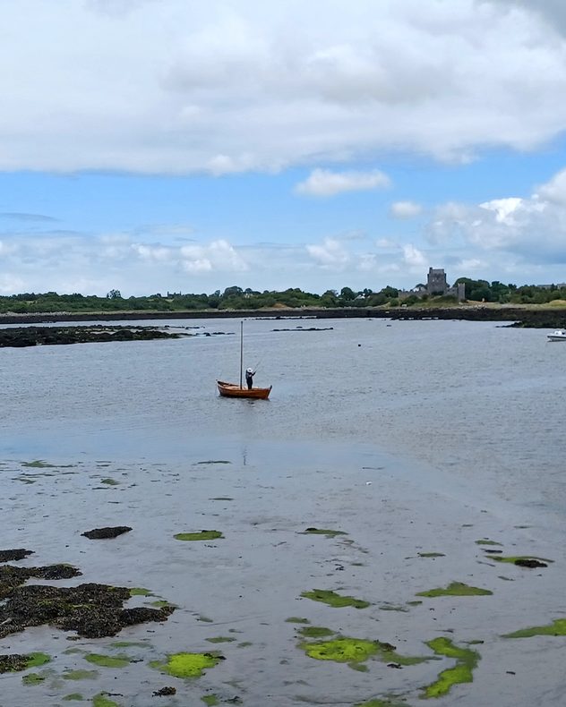 Small wooden sailboat with one person aboard on a calm coastal bay at low tide, with exposed rocks, green seaweed, and a distant stone tower beneath broken clouds.