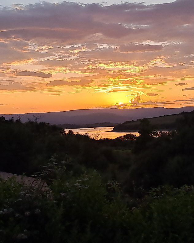 Golden sun setting behind distant hills beneath layered clouds, with orange light reflecting across a winding body of water and dark vegetation in the foreground.