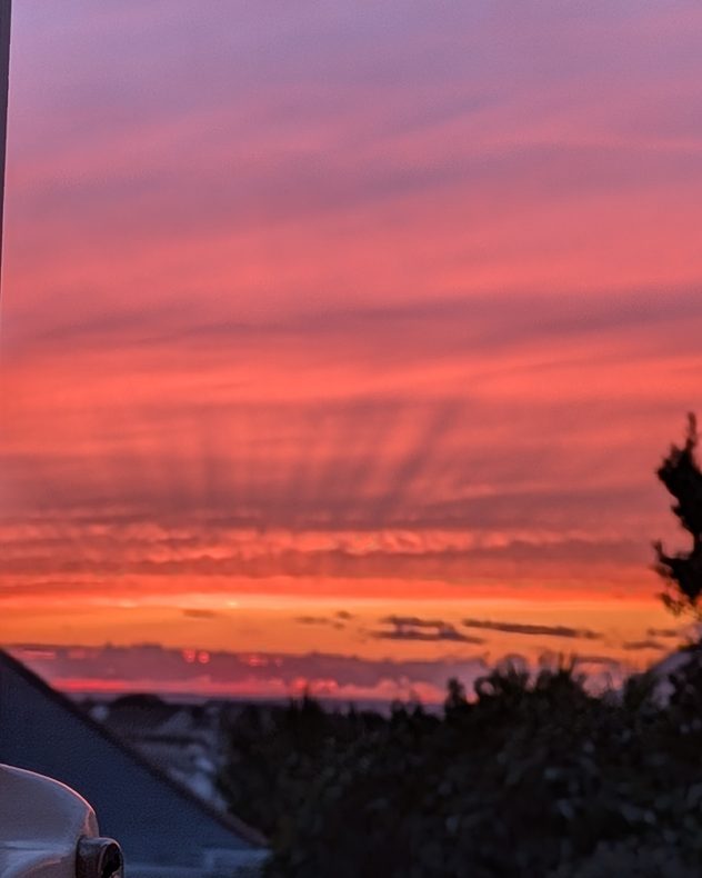 Vivid pink, red and orange sunset clouds layered above silhouetted rooftops and trees, with darker streaks radiating through the glowing evening sky.