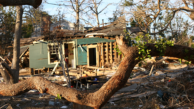 Ein Haus ist schwer beschädigt, Wände und Dach eingestürzt, Trümmer liegen verstreut. Im Vordergrund ein umgestürzter Baum.