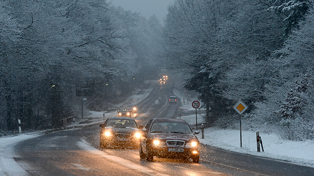 Autos fahren bei Schneefall auf verschneiter Landstraße mit eingeschaltetem Licht.