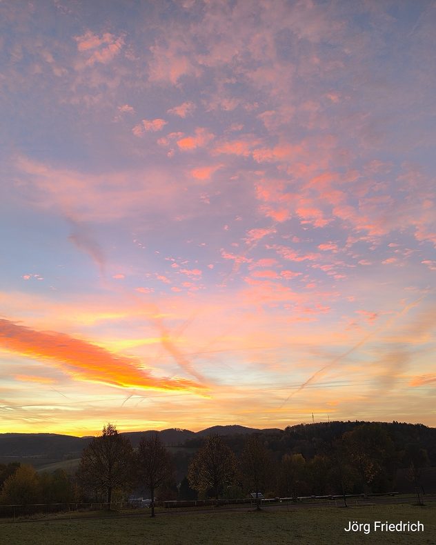 Farbenfroher Sonnenaufgang über einer Hügellandschaft mit Bäumen im Vordergrund.
