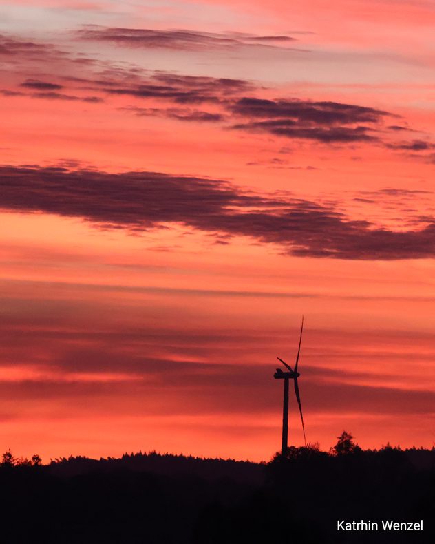 Windrad vor rotem Morgenhimmel mit leuchtenden Wolkenschichten.