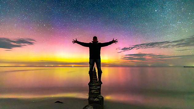 Silhouette einer Person auf Holzpfählen im Meer, umgeben von farbenfrohen Polarlichtern, die sich im Wasser spiegeln.