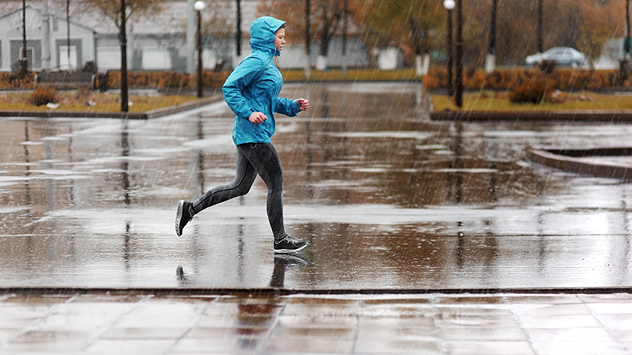 Person joggt im strömenden Regen über eine nasse, spiegelnde Fläche in herbstlicher Stadtumgebung – Kleidung wetterfest, Umgebung menschenleer.
