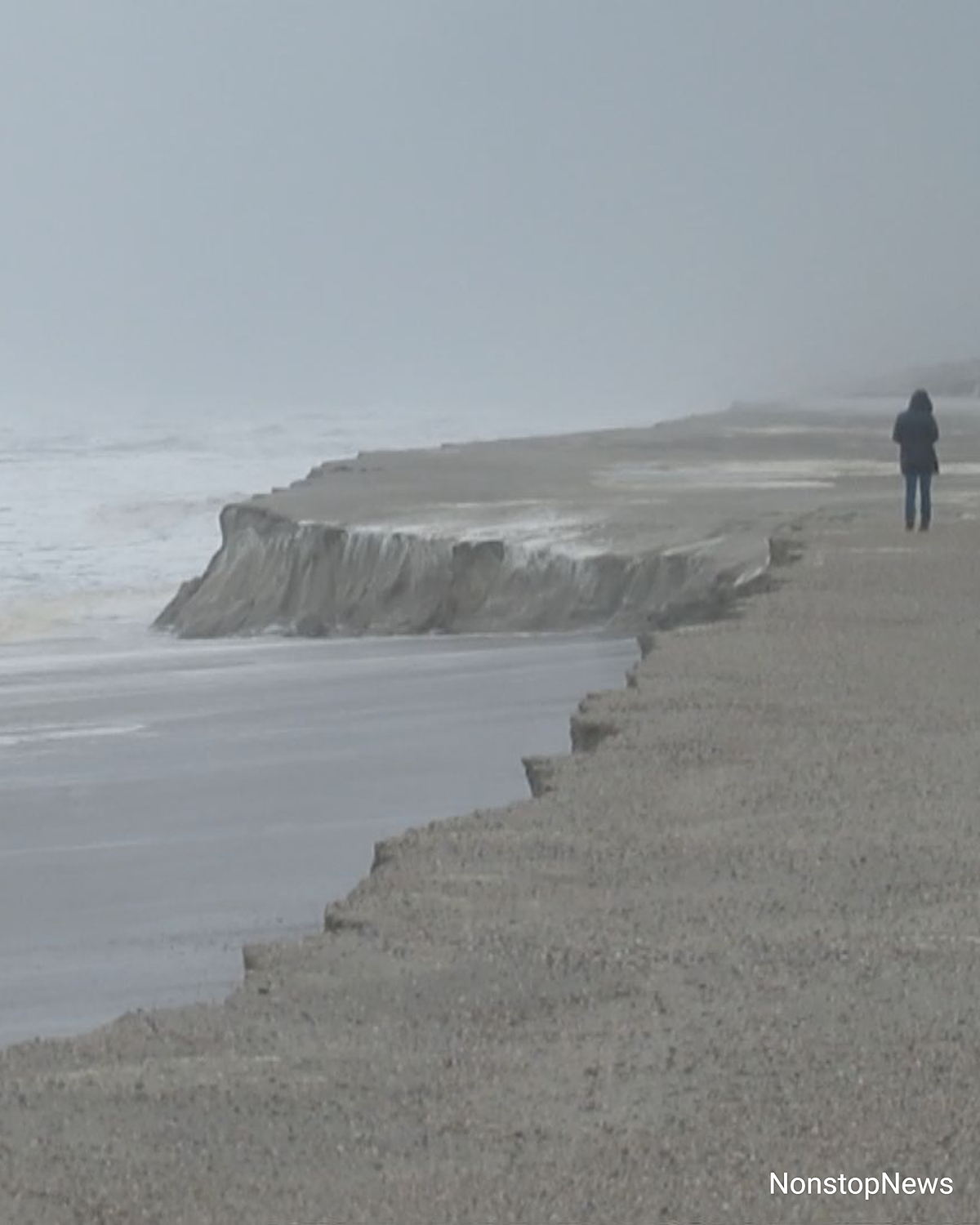 Strand von Langeoog mit steiler Sandabbruchkante nach einer Sturmflut, graues Wetter und eine Person im Hintergrund.