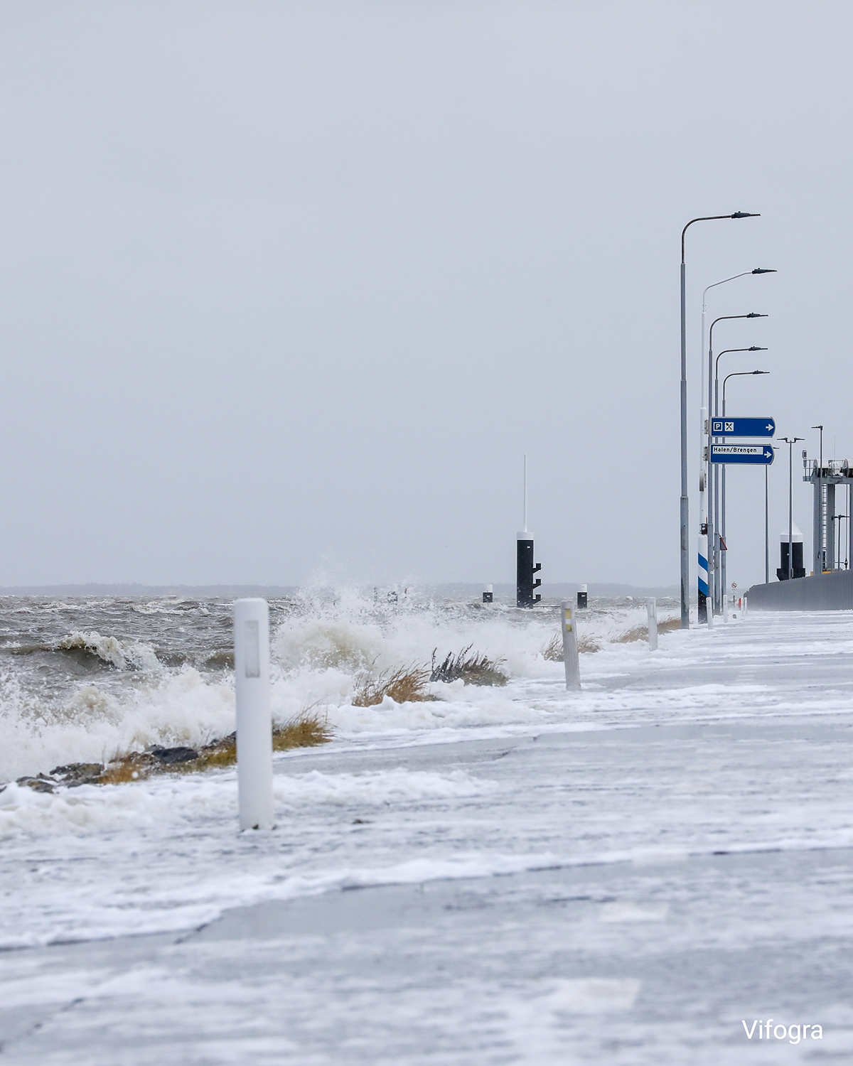 Überspülte Küstenstraße bei Holwert, Wellen schlagen über die Fahrbahn, Schaum und stürmisches Meer unter grauem Himmel.