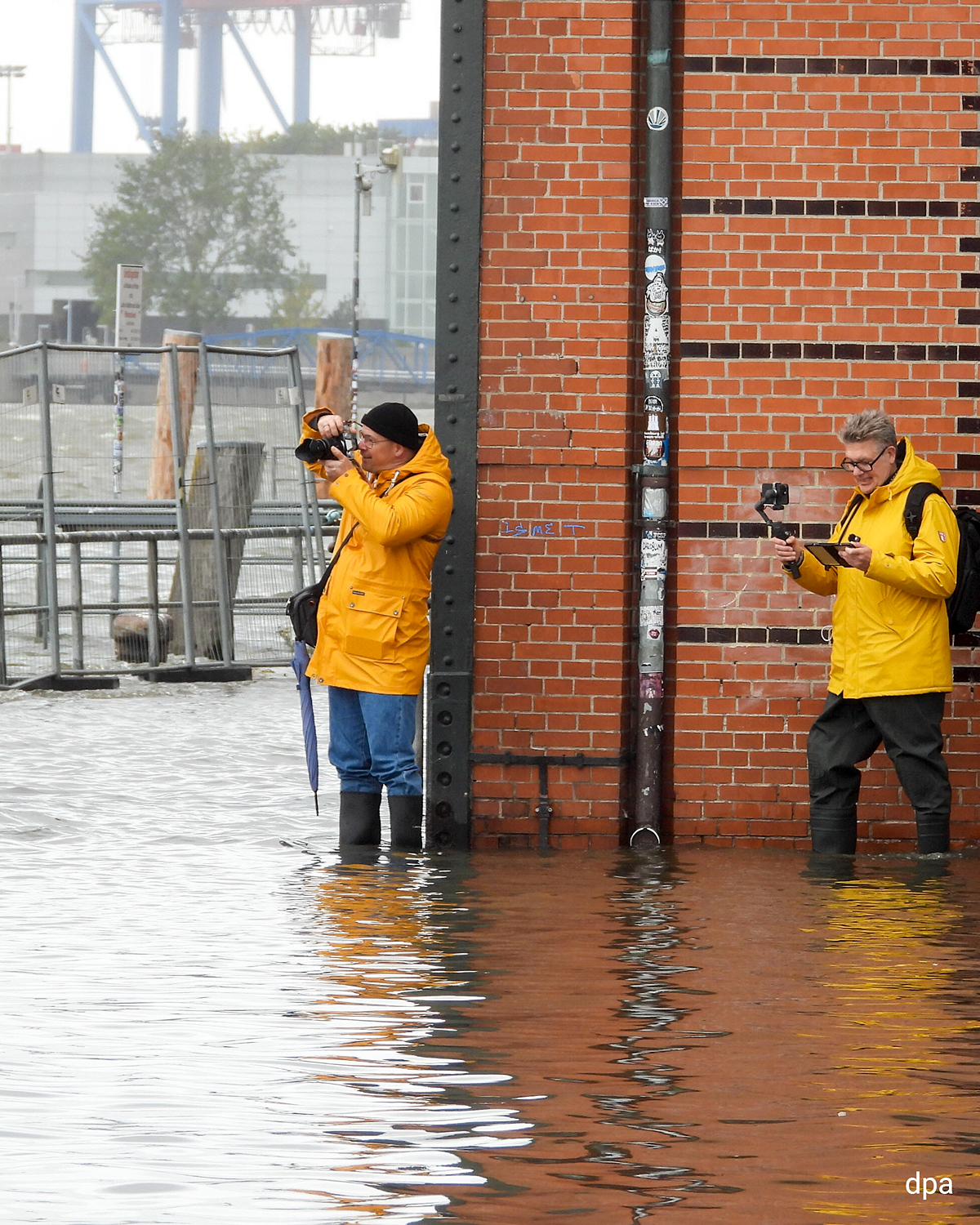 Zwei Personen in gelben Regenjacken stehen im überfluteten Bereich des Hamburger Fischmarkts und fotografieren das Hochwasser.