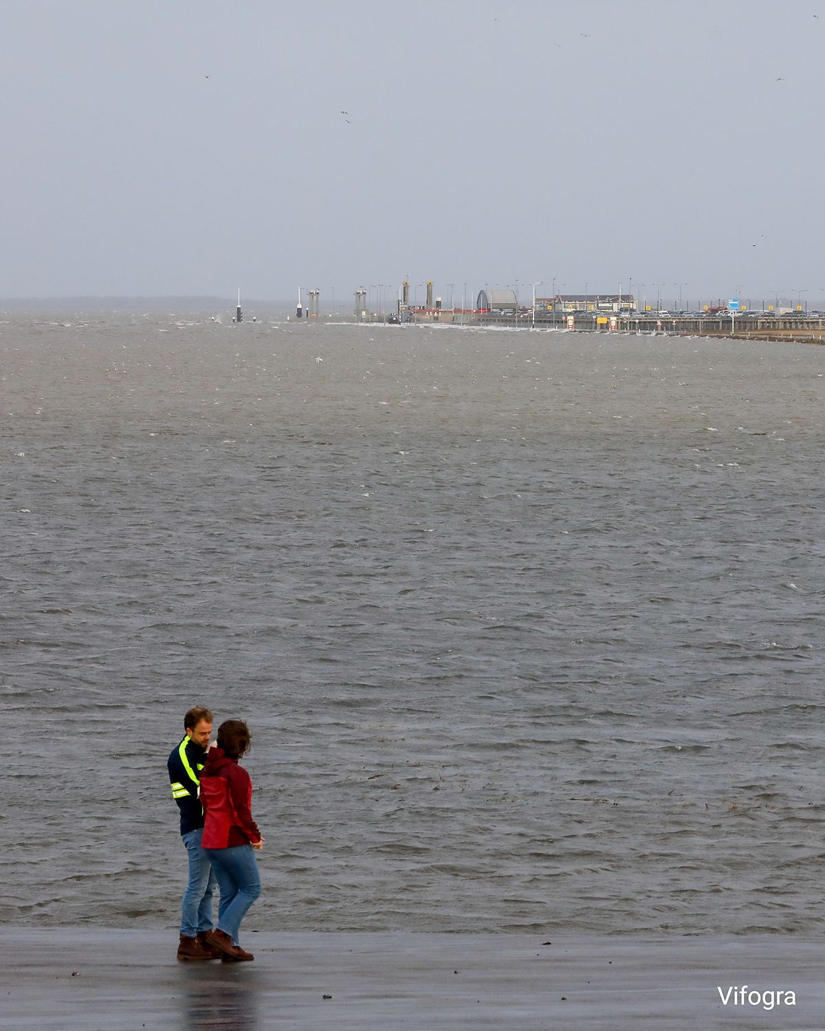Zwei Personen gehen am Deich entlang, im Hintergrund der überflutete Fährhafen von Holwert bei Sturmflut und grauem Himmel.