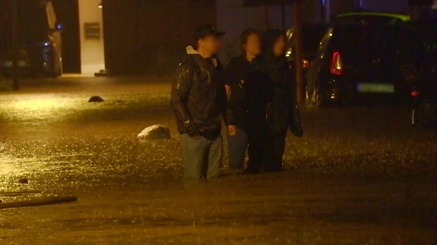 Menschen stehen nachts in einer überfluteten Straße, umgeben von Autos.