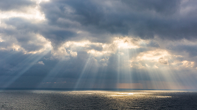 Снимка на която виждаме как слънчевите лъчи пробиват през облаците над морето. В далечината се виждат и дъждовни завеси