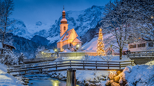 Winterliche Alpenlandschaft mit schneebedecktem Dorf, beleuchteter Kirche und festlich geschmücktem Weihnachtsbaum an einem Bach bei Abenddämmerung.