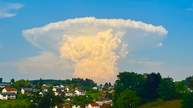 Eine gewaltige weiße Wolke türmt sich über einem Dorf mit roten Dächern, klarer Himmel im Hintergrund.