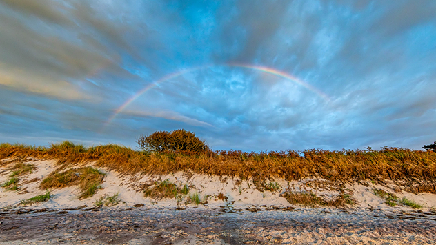 Ein Regenbogen spannt sich über Dünen mit Grasbewuchs, im Vordergrund liegt ein Sandstrand.