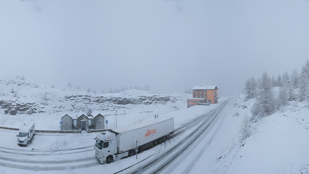 Zwei Lastwagen fahren auf einer schneebedeckten Passstraße bei Nebel. Die Umgebung ist vollständig verschneit, im Hintergrund steht ein Haus. Die Sichtverhältnisse sind winterlich eingeschränkt.