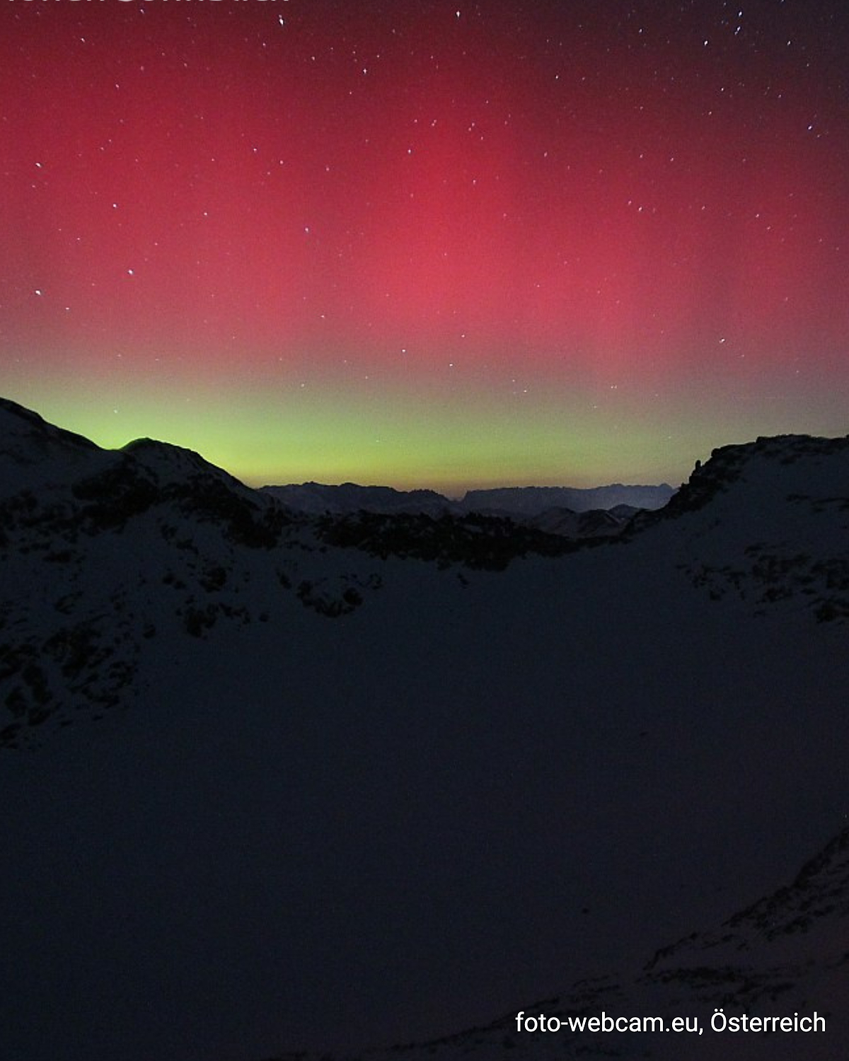 Rotes und grünes Polarlicht über verschneiten Alpen bei Nacht.