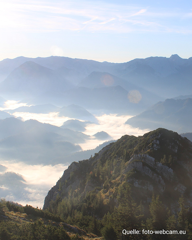 Blick über bewaldete und felsige Berggipfel auf ein nebelgefülltes Tal. Im Hintergrund staffeln sich mehrere Gebirgsketten im Dunst, während die tief stehende Sonne die Landschaft in warmes Licht taucht. Ist dieses Gespräch bisher hilfreich?