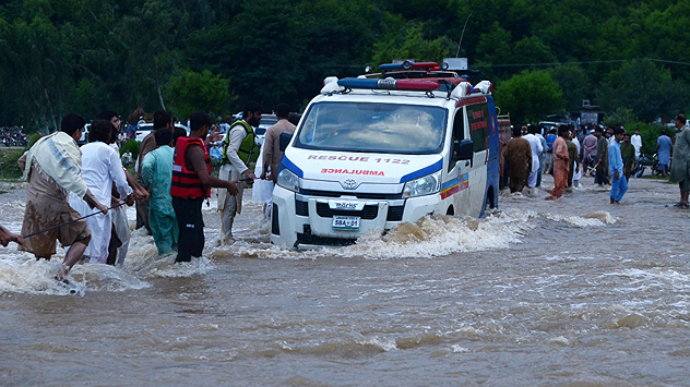 Rettungskräfte begleiten Krankenwagen durch überflutete Straße in Pakistan, zahlreiche Menschen im Wasser, dichter Wald im Hintergrund.