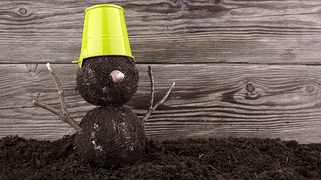 Ein Schneemann aus Erde statt Schnee steht vor einer Holzwand. Er besteht aus zwei Erdbällen, hat Ärmchen aus Zweigen, eine Schneckenschale als Nase und trägt einen umgedrehten grünen Blumentopf als Hut – humorvolle Szene bei mildem Winterwetter.