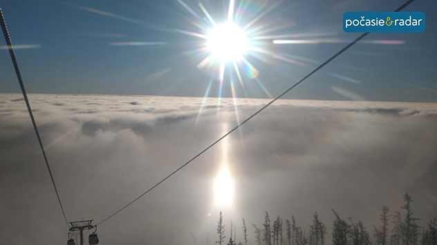 Hmla nad ktorou svieti Slnko. Fotografia je fotená z kabínkovej lanovky, preto je vidno aj laná a dve kabínky v spodnej časti obrázku.