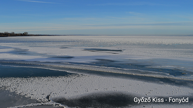 Velika, djelomično zatvorena ledena površina na Balatonu. U prvom planu vide se slomljene sante leda i tamna voda. Ravna brda leže na horizontu pod vedrim nebom.