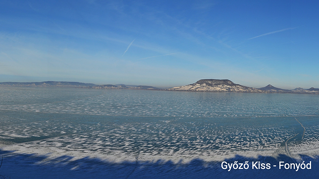 Jezero Balaton je uglavnom zaleđeno s ledenim formacijama na površini. U pozadini se vide brda i suprotna obala. Vedro nebo iznad zimskog jezerskog krajolika.