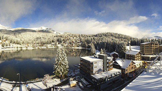 © roundshot.com Verschneite Ortschaft an einem Bergsee mit schneebedeckten Wäldern und klarer Winterluft.