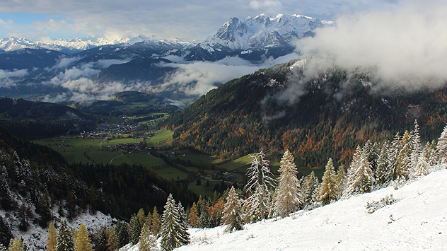 © foto-webcam.eu Vordergrund: Verschneite Nadelbäume an einem Berghang; Hintergrund: Das darunterliegende grüne Tal mit verfärbten Laubbäumen
