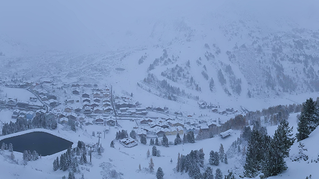 Ein Bergdorf im Morgengrauen ist verschneit. Im Vordergrund sind Nadelbäume und ein kleiner See, im Hintergrund Berge.