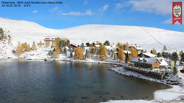 © foto-webcam.eu Eine Siedlung in den Bergen mit einem See im Vordergrund ist mit Schnee bedeckt. Die Sonne scheint und der Himmel ist blau
