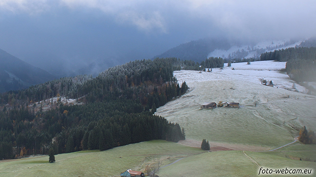 © foto-webcam.eu Teilweise verschneite Wiesen und Wälder in hügeliger Berglandschaft unter tiefen Wolken.
