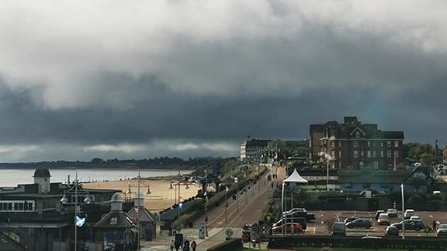 A coastal town under dark, heavy clouds, with a beach and promenade lit by a patch of sunlight in the foreground and a stormy sky moving in from the sea.