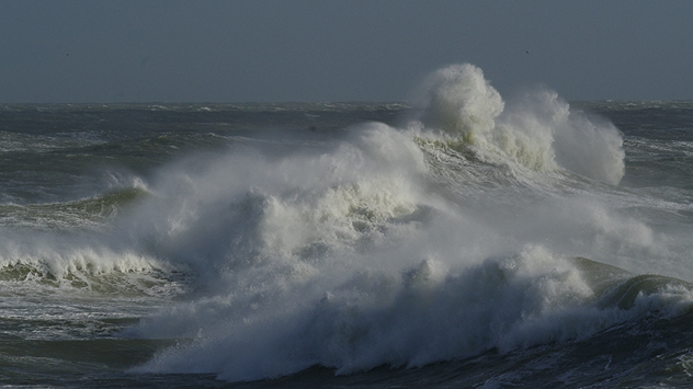 Large, powerful waves crash and churn in a dark, stormy sea, with white spray and foam whipped up by strong winds under a grey sky.