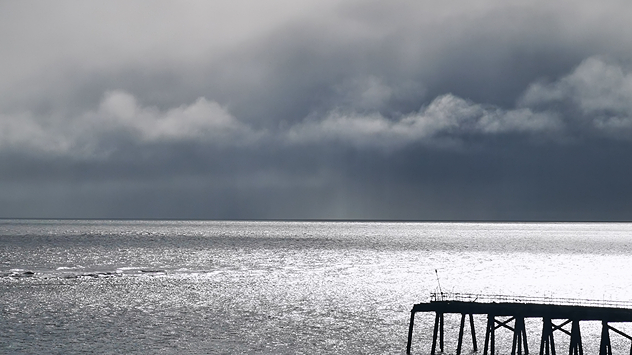 Dark storm clouds hang low over a silvery, sunlit sea, with light rain visible on the horizon and part of a pier extending into the water in the foreground.