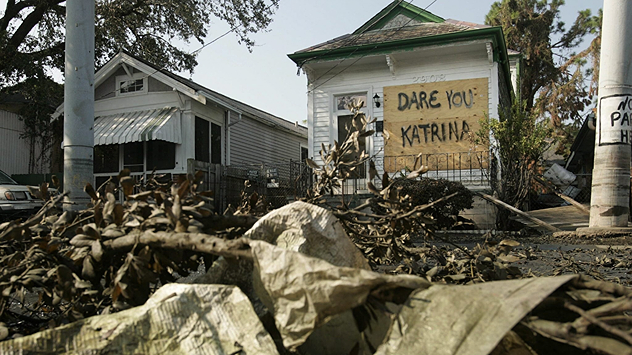 Et hus med et håndskrevet skilt med teksten „Dare You Katrina“ mellem ødelagte grene og murbrokker.