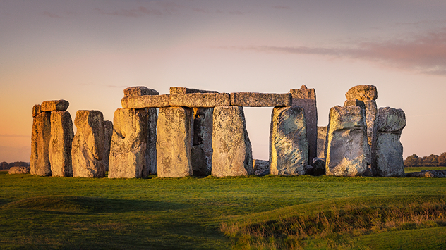 Stonehenge standing on green grass in warm golden-hour light beneath a pale pink and blue evening sky.
