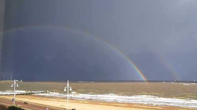 A double rainbow arcs over the sea, with dark grey storm clouds in the background and waves breaking onto a sandy beach in the foreground.