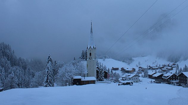Verschneite Kirche in nebliger Winterlandschaft mit Häusern und Bäumen. Ruhige Szenerie im dichten Neuschnee. (c) foto-webcam.eu