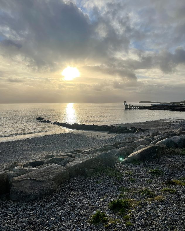Soft sunrise over a calm sea with golden light reflecting on the water, rocky foreground, a narrow breakwater leading offshore, and a small pier structure on the right under layered clouds.