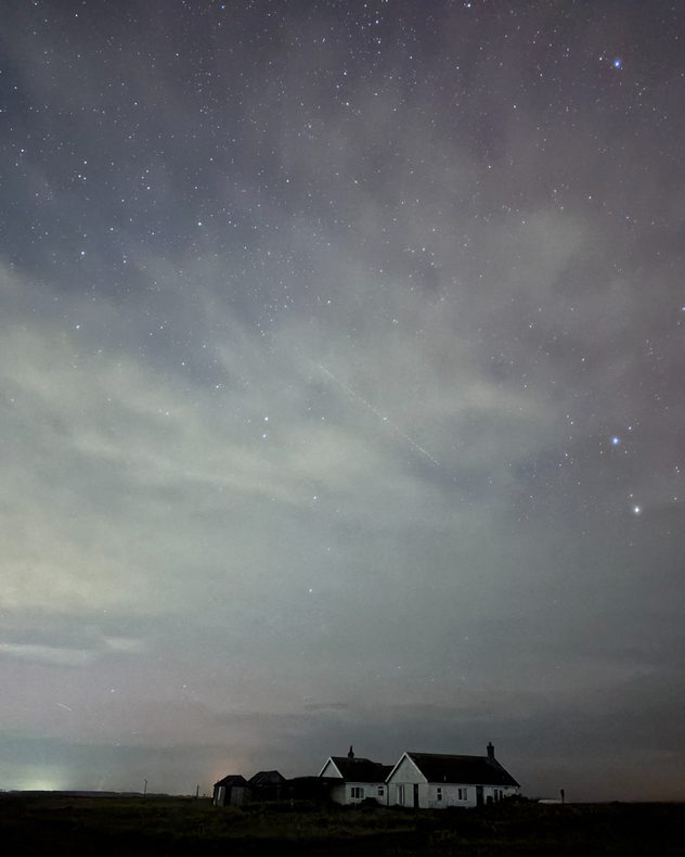 Night scene with a star-filled sky above a small white cottage and outbuildings on open land, thin clouds drifting across the stars and a faint aurora.
