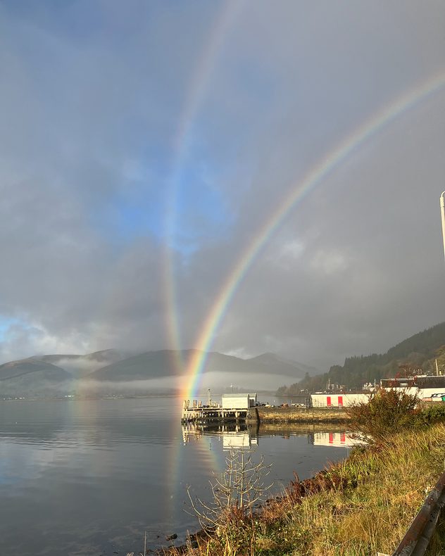 Double rainbow arching above a calm lakeside with mountains in the background, bright reflections on the water, a small pier and buildings to the right, and low mist along the slopes.