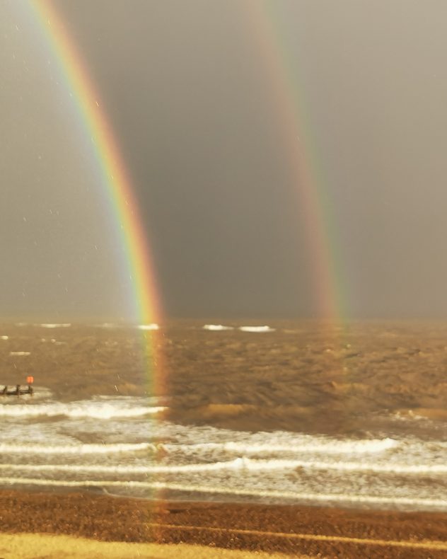 Double rainbow arcs over a rough, wind-stirred North Sea with white waves breaking towards a sandy shore under a dark grey sky.