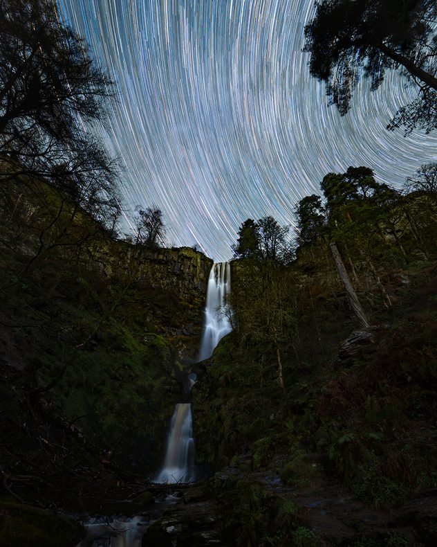 Long-exposure night scene of a tall, tiered waterfall flowing through a forested gorge, with bright circular star trails sweeping across the sky above.